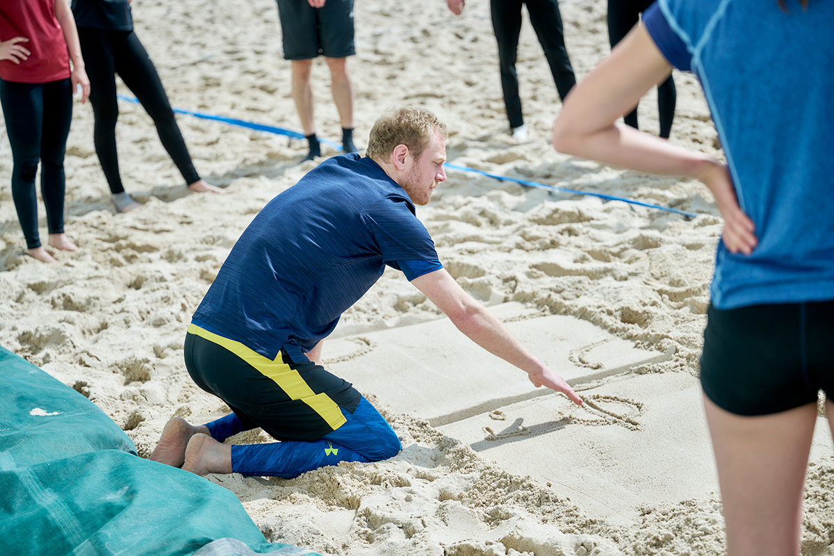 Ein Lehrer erklärt eine Regel im Beachvolleyball und zeichnet sie dazu im Sand.